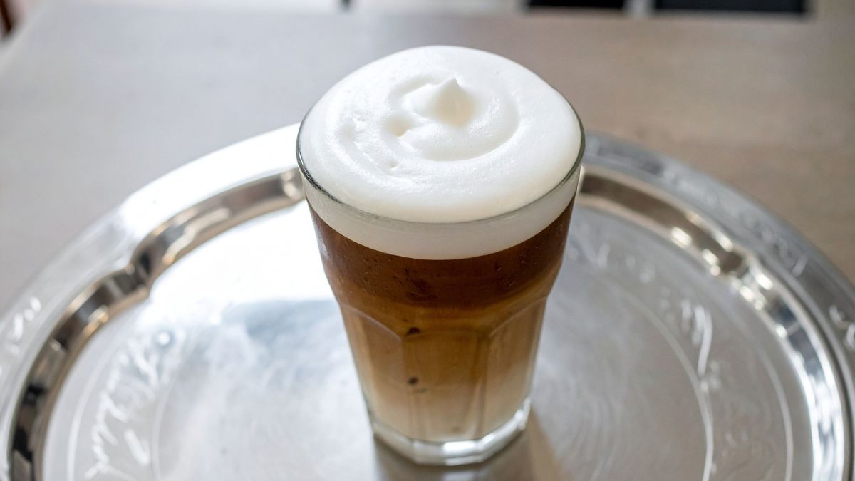 Egy pohár kávé, tetején sűrű, krémes tejhab, fényes tálcán.