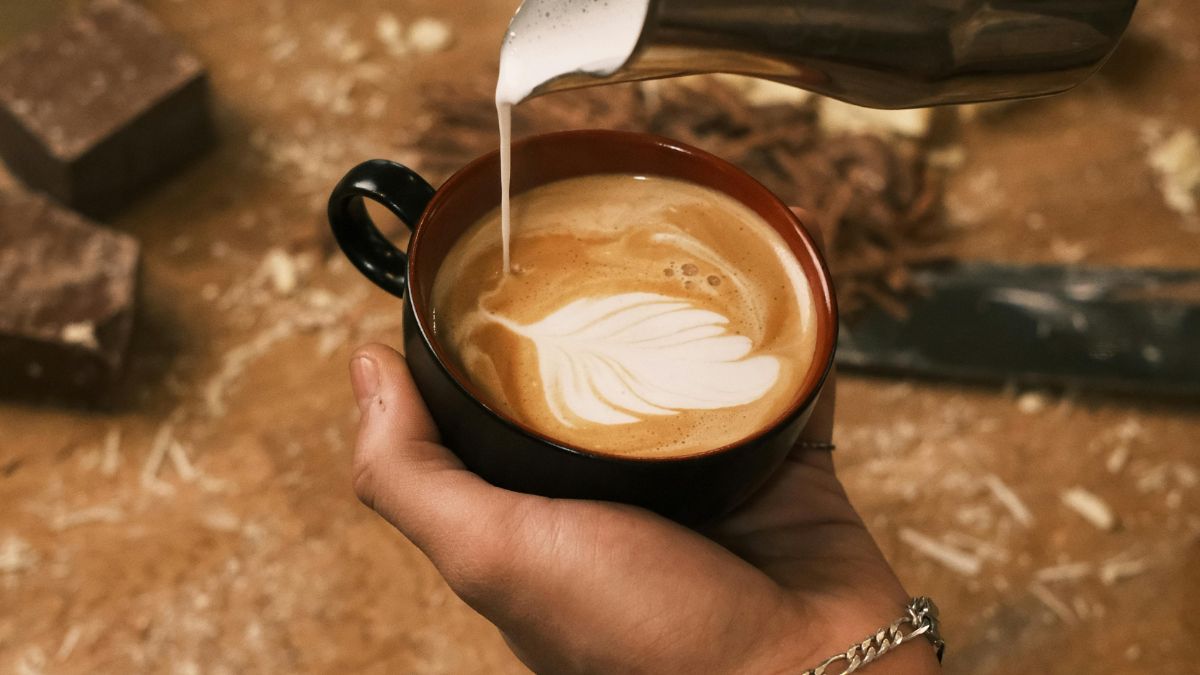 Egy kéz egy fekete bögrét tart, amelyben krémes kávé és tejhab látható.
